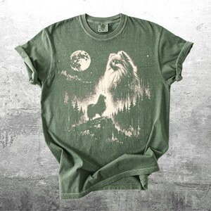 May include: Green t-shirt featuring a graphic of a dog howling at the moon in a forest setting. The design is in shades of white and tan against the green fabric. The shirt has a classic crew neck and short sleeves.