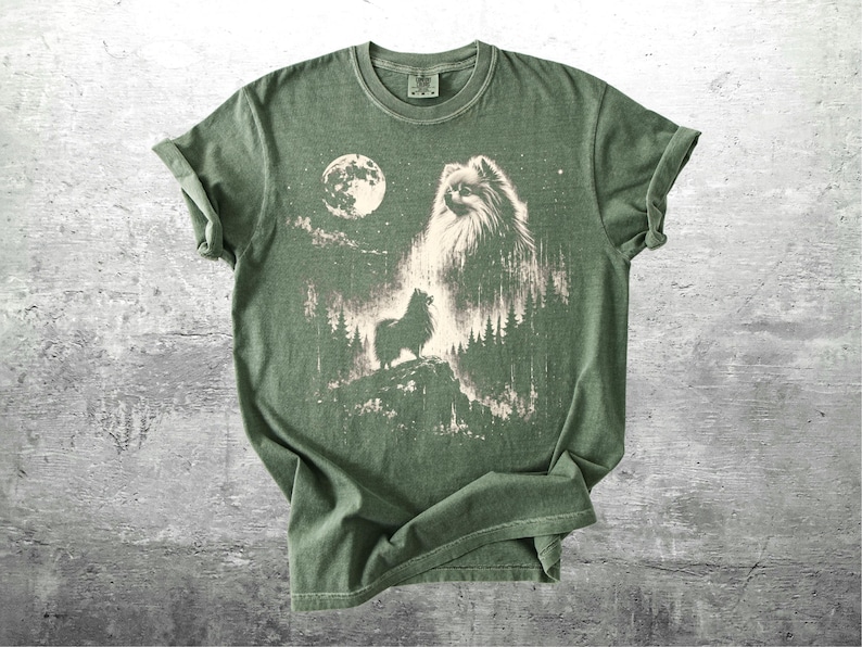 May include: Green t-shirt featuring a graphic of a dog howling at the moon in a forest setting. The design is in shades of white and tan against the green fabric. The shirt has a classic crew neck and short sleeves.