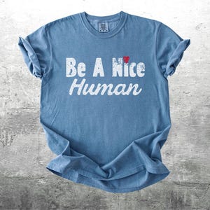 K&ouml;nnte beinhalten: Ein blaues T-Shirt mit den Worten "Be A Nice Human" in abgenutzten wei&szlig;en Buchstaben. Ein kleines rotes Herz befindet sich &uuml;ber dem Wort "Nice". Das T-Shirt ist gefaltet und liegt auf einer grauen Oberfl&auml;che.