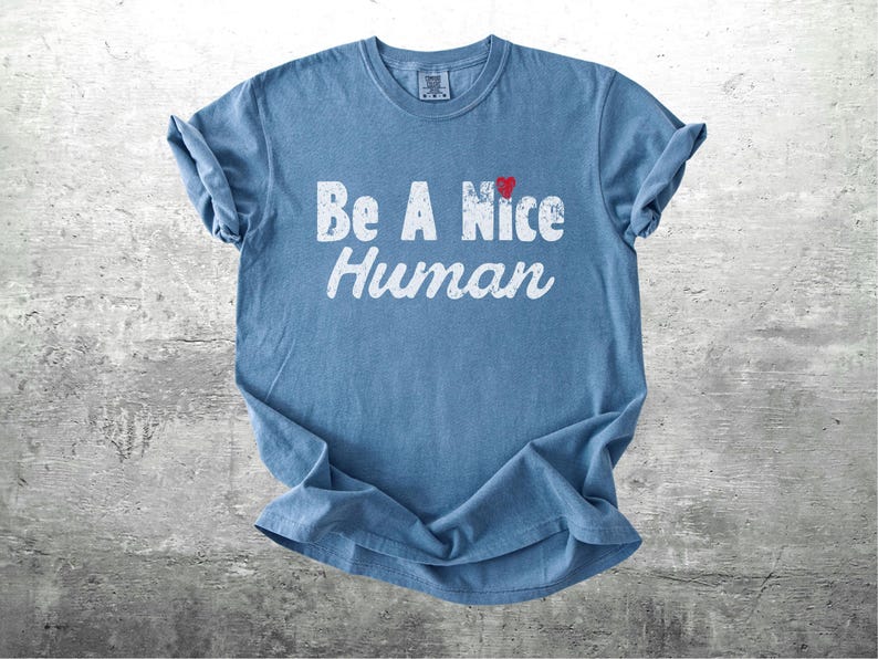 K&ouml;nnte beinhalten: Ein blaues T-Shirt mit den Worten "Be A Nice Human" in abgenutzten wei&szlig;en Buchstaben. Ein kleines rotes Herz befindet sich &uuml;ber dem Wort "Nice". Das T-Shirt ist gefaltet und liegt auf einer grauen Oberfl&auml;che.