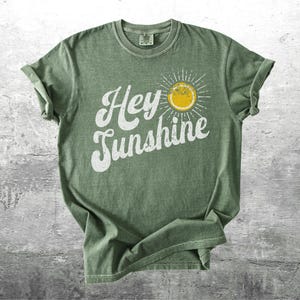May include: A sage green t-shirt with the words "Hey Sunshine" in white script, accompanied by a yellow sun graphic with radiating lines. The shirt has rolled-up sleeves and a vintage, worn look.