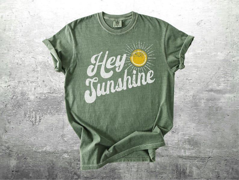 May include: A sage green t-shirt with the words "Hey Sunshine" in white script, accompanied by a yellow sun graphic with radiating lines. The shirt has rolled-up sleeves and a vintage, worn look.