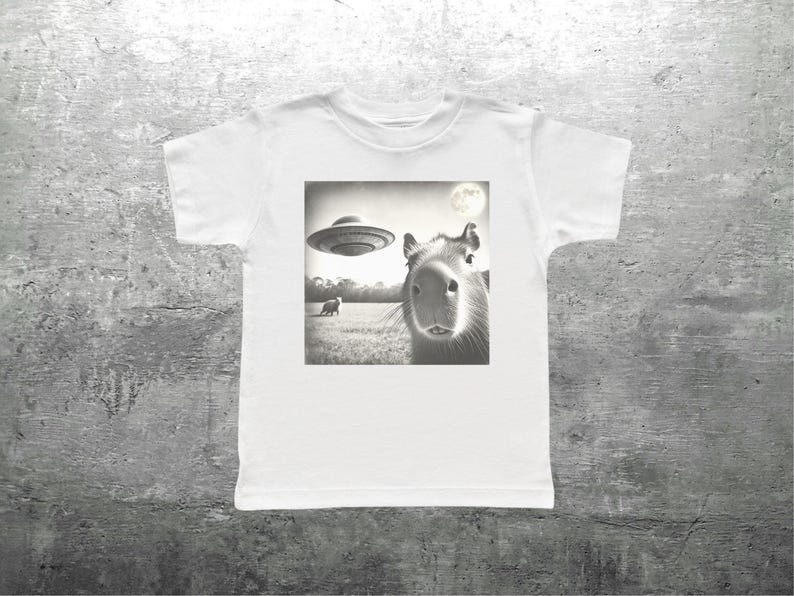 May include: A white t-shirt with a black and white image of a capybara looking up at a flying saucer. The image also shows a small animal in the background.