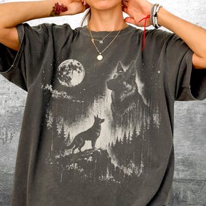 May include: Dark gray t-shirt featuring a wolf graphic under a full moon. The design includes a silhouette of a wolf on a rocky outcrop, with a larger wolf head and forest imagery.