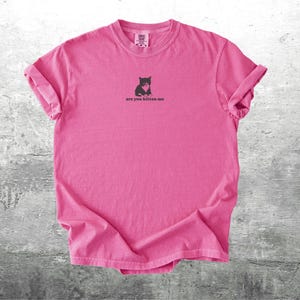 May include: A bright pink t-shirt with rolled-up sleeves. The shirt features a small graphic of a black cat holding a heart, with the text "are you kitten me" printed below the graphic. The shirt is made of a soft material.