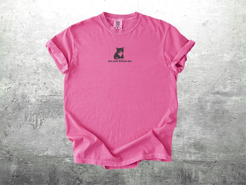 May include: A bright pink t-shirt with rolled-up sleeves. The shirt features a small graphic of a black cat holding a heart, with the text "are you kitten me" printed below the graphic. The shirt is made of a soft material.
