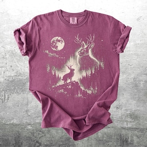 May include: A maroon t-shirt featuring a graphic of a deer-rabbit hybrid with antlers, set against a forest and a full moon. The design is in a faded, vintage style. The shirt is folded, showing the texture of the fabric.