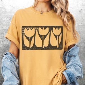 May include: Mustard yellow t-shirt featuring a black rectangular graphic of three stylized tulip flowers. The flowers have a simple, blocky design. The shirt has rolled-up sleeves and is paired with a denim jacket.