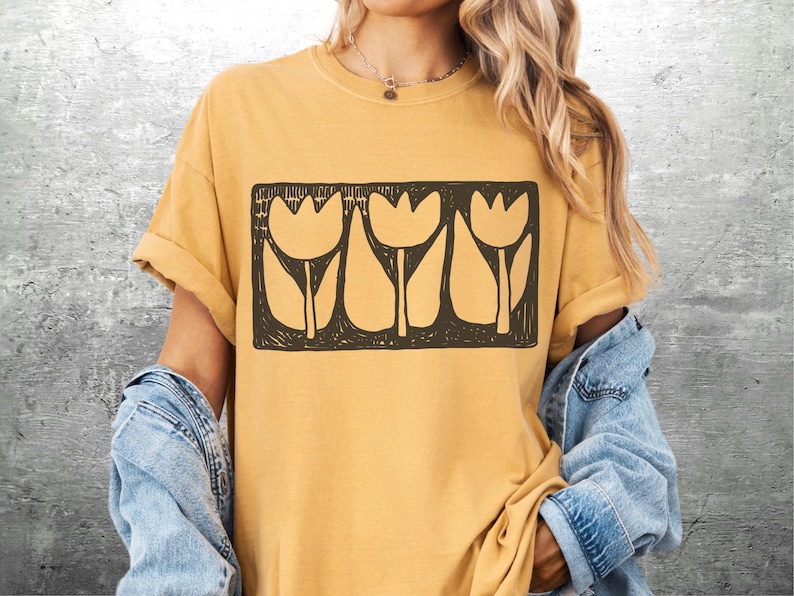 May include: Mustard yellow t-shirt featuring a black rectangular graphic of three stylized tulip flowers. The flowers have a simple, blocky design. The shirt has rolled-up sleeves and is paired with a denim jacket.