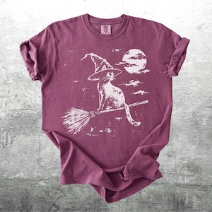 May include: A maroon short-sleeve t-shirt featuring a white graphic print of a cat wearing a witch hat and sitting on a broomstick against a full moon and clouds. The shirt has a crew neckline and rolled sleeves, with a distressed texture.