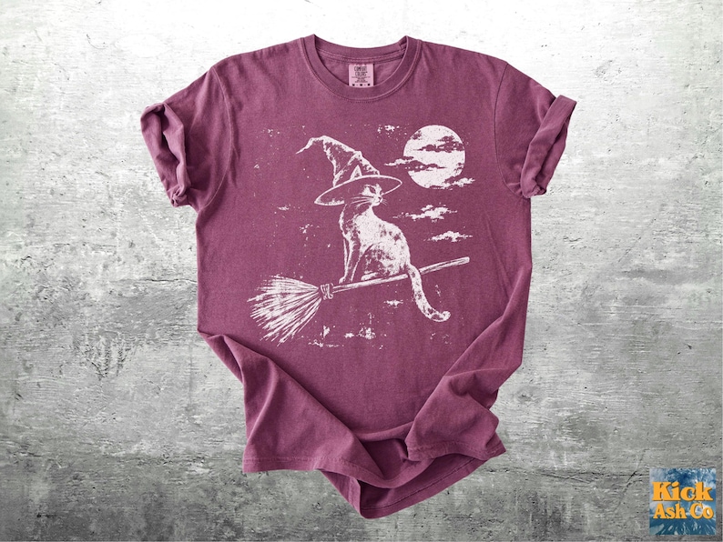 May include: A maroon short-sleeve t-shirt featuring a white graphic print of a cat wearing a witch hat and sitting on a broomstick against a full moon and clouds. The shirt has a crew neckline and rolled sleeves, with a distressed texture.