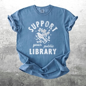 May include: A blue t-shirt with the words "SUPPORT your public LIBRARY" in white, along with a graphic of a bird on a branch with flowers. The shirt is folded and lies on a gray surface.