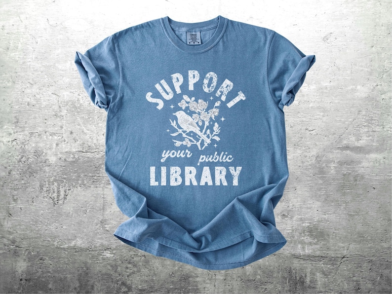 May include: A blue t-shirt with the words "SUPPORT your public LIBRARY" in white, along with a graphic of a bird on a branch with flowers. The shirt is folded and lies on a gray surface.
