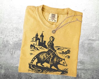 May include: Mustard yellow t-shirt with a black graphic of a knight riding a possum in front of a castle. A silver necklace with a circular pendant rests on the shirt. The shirt has a Comfort Colors tag.