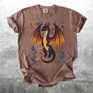 May include: A brown t-shirt with a black dragon with orange and yellow wings and floral details printed on the front.