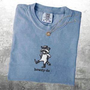 May include: A light blue Comfort Colors t-shirt with a graphic of a raccoon wearing a cowboy hat and boots, with the text "howdy-do" below. A gold necklace with a pendant is draped across the shirt.