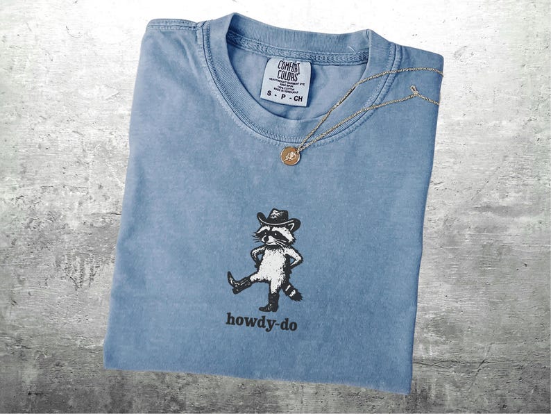 May include: A light blue Comfort Colors t-shirt with a graphic of a raccoon wearing a cowboy hat and boots, with the text "howdy-do" below. A gold necklace with a pendant is draped across the shirt.
