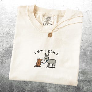 May include: Off-white t-shirt featuring the text "I don't give a" above a cartoon of a mouse and donkey. The mouse holds a leash attached to the donkey. A gold necklace with a pendant is draped across the shirt.
