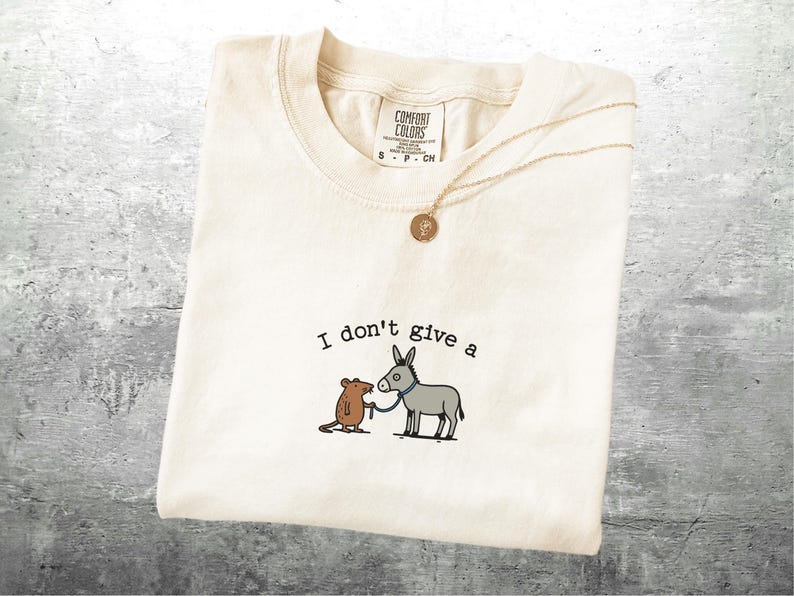 May include: Off-white t-shirt featuring the text "I don't give a" above a cartoon of a mouse and donkey. The mouse holds a leash attached to the donkey. A gold necklace with a pendant is draped across the shirt.
