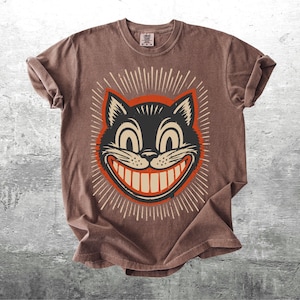 Op de afbeelding: Een bruin t-shirt met korte mouwen en een afbeelding van een zwarte kattenkop met een brede, tandige grijns. De kat is omlijst door een rode cirkel en witte stralende lijnen. Het t-shirt heeft een ronde hals en een label in de nek.