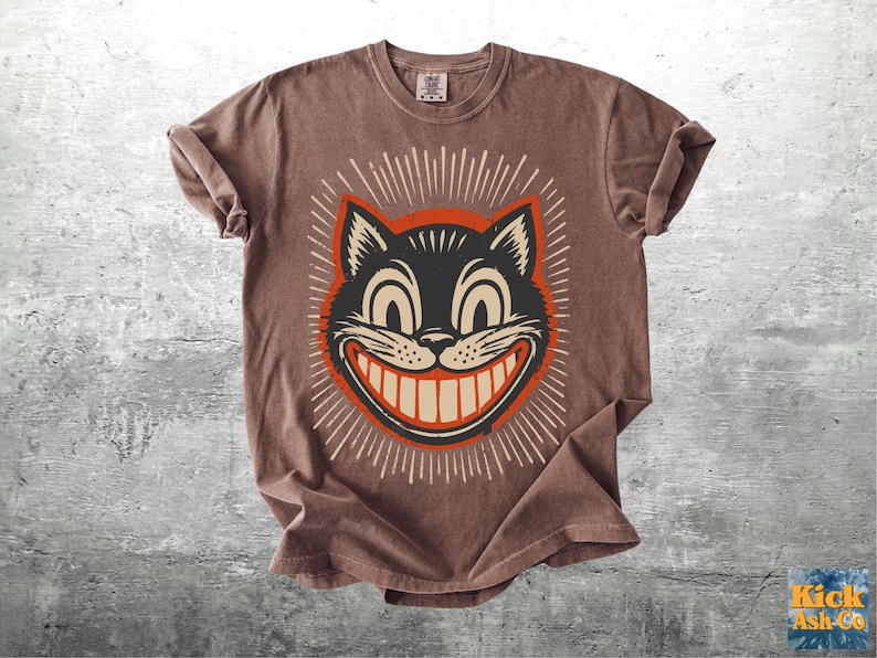 Op de afbeelding: Een bruin t-shirt met korte mouwen en een afbeelding van een zwarte kattenkop met een brede, tandige grijns. De kat is omlijst door een rode cirkel en witte stralende lijnen. Het t-shirt heeft een ronde hals en een label in de nek.