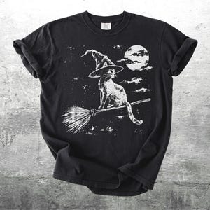 May include: A black short-sleeve t-shirt featuring a white graphic print of a cat wearing a pointed witch hat while sitting on a broomstick. The design includes a full moon and clouds in the background, rendered in a distressed, textured style.