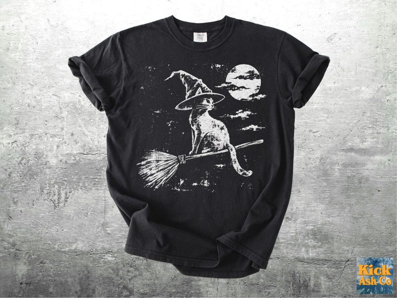 May include: A black short-sleeve t-shirt featuring a white graphic print of a cat wearing a pointed witch hat while sitting on a broomstick. The design includes a full moon and clouds in the background, rendered in a distressed, textured style.