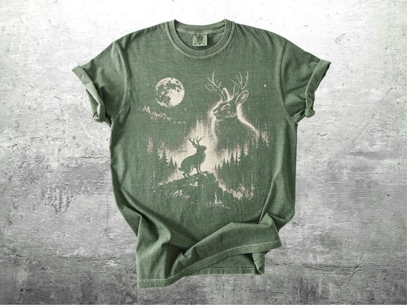 May include: Green t-shirt featuring a graphic of a jackalope with antlers, set against a forest and moon backdrop. The design is in a faded, vintage style. The shirt is made of soft material and has a classic crew neck.