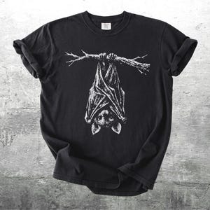 Op de afbeelding: Zwart t-shirt met een witte illustratie van een vleermuis die ondersteboven aan een tak hangt. De vleermuis heeft gedetailleerde vleugels en een gerichte blik. Het shirt is gemaakt van een zacht materiaal.