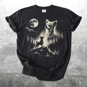 May include: Black t-shirt featuring a graphic of a Chihuahua dog under a full moon in a forest setting. The design includes a large dog head and a smaller dog standing on a hill. The artwork is in shades of white and tan.