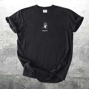 May include: Black t-shirt with a small white graphic of a raccoon wearing a cowboy hat. The text "howdy-do" is printed below the raccoon. The shirt is made of a soft material and has rolled-up sleeves.