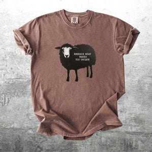 Puede incluir: Camiseta marr&oacute;n con un dise&ntilde;o de oveja negra y el texto "Embrace What Makes You Unique". La camiseta tiene cuello redondo y mangas cortas. La tela parece suave y ligeramente arrugada.