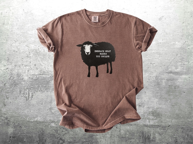 Puede incluir: Camiseta marr&oacute;n con un dise&ntilde;o de oveja negra y el texto "Embrace What Makes You Unique". La camiseta tiene cuello redondo y mangas cortas. La tela parece suave y ligeramente arrugada.