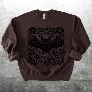 May include: A dark brown sweatshirt featuring a black bat design surrounded by flowers and leaves. The bat has outstretched wings and is centered on the front of the long-sleeved top. The sleeves are rolled up.