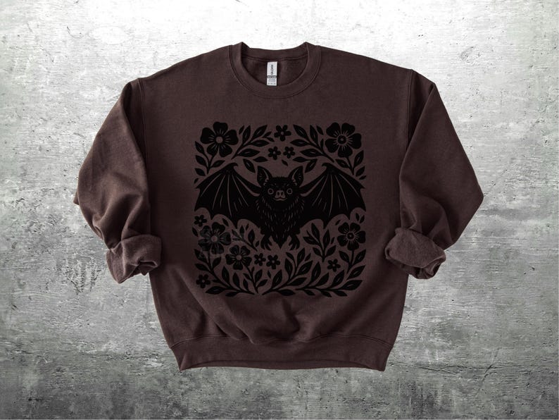 May include: A dark brown sweatshirt featuring a black bat design surrounded by flowers and leaves. The bat has outstretched wings and is centered on the front of the long-sleeved top. The sleeves are rolled up.