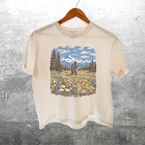 Puede incluir: Camiseta color crema con un gr&aacute;fico de un Bigfoot en un prado con monta&ntilde;as y &aacute;rboles. El dise&ntilde;o incluye cielos azules y flores coloridas. La camiseta est&aacute; colgada en una percha de madera.