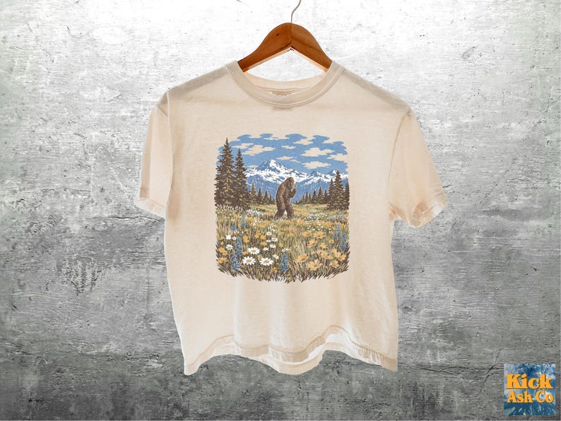 Puede incluir: Camiseta color crema con un gr&aacute;fico de un Bigfoot en un prado con monta&ntilde;as y &aacute;rboles. El dise&ntilde;o incluye cielos azules y flores coloridas. La camiseta est&aacute; colgada en una percha de madera.