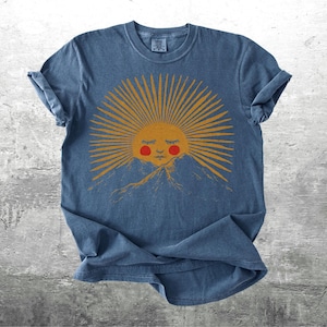 K&ouml;nnte beinhalten: Blaues T-Shirt mit einer Grafik einer l&auml;chelnden Sonne mit orangefarbenen Strahlen und roten Wangen, vor einer Bergkette. Das Design ist auf einem weichen, l&auml;ssigen T-Shirt gedruckt.