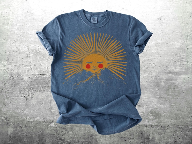 K&ouml;nnte beinhalten: Blaues T-Shirt mit einer Grafik einer l&auml;chelnden Sonne mit orangefarbenen Strahlen und roten Wangen, vor einer Bergkette. Das Design ist auf einem weichen, l&auml;ssigen T-Shirt gedruckt.