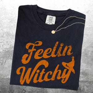 K&ouml;nnte beinhalten: Ein marineblaues T-Shirt mit dem Schriftzug "Feelin Witchy" in orangefarbenen, verwaschenen Buchstaben und einer Hexenhut-Grafik. Eine goldene Halskette mit rundem Anh&auml;nger liegt auf dem Shirt. Das Shirt liegt auf einer grauen Oberfl&auml;che.