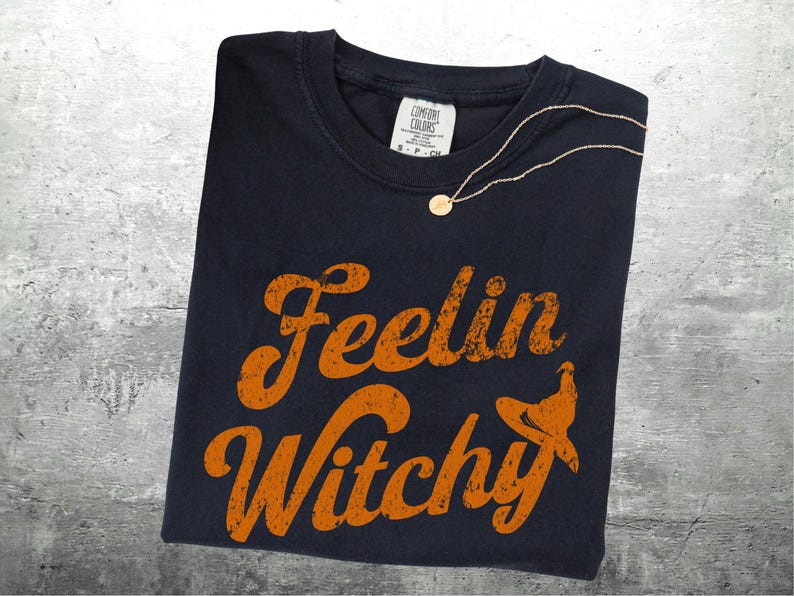 K&ouml;nnte beinhalten: Ein marineblaues T-Shirt mit dem Schriftzug "Feelin Witchy" in orangefarbenen, verwaschenen Buchstaben und einer Hexenhut-Grafik. Eine goldene Halskette mit rundem Anh&auml;nger liegt auf dem Shirt. Das Shirt liegt auf einer grauen Oberfl&auml;che.