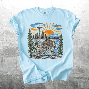 May include: A light blue t-shirt featuring a graphic print of a raccoon walking in front of a lake, pine trees, a city skyline with the Space Needle, and a rising sun. The text Kick Ash Co is printed in the bottom right corner of the image.