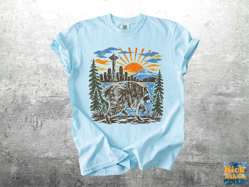May include: A light blue t-shirt featuring a graphic print of a raccoon walking in front of a lake, pine trees, a city skyline with the Space Needle, and a rising sun. The text Kick Ash Co is printed in the bottom right corner of the image.