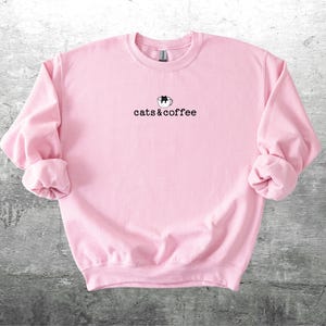 May include: A light pink sweatshirt with the text "cats & coffee" in a black, simple font. Above the text is a small graphic of a cat in a coffee cup. The sweatshirt has long sleeves with cuffed wrists.