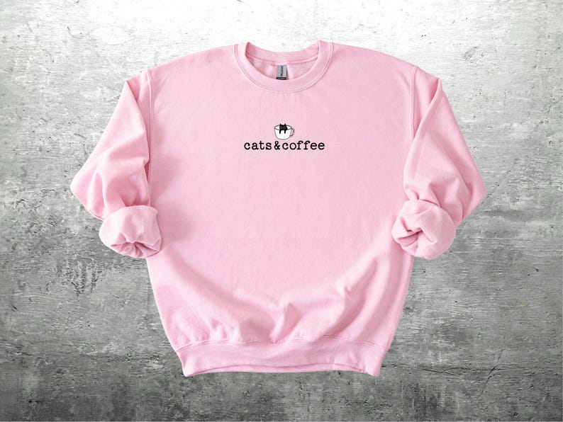 May include: A light pink sweatshirt with the text "cats & coffee" in a black, simple font. Above the text is a small graphic of a cat in a coffee cup. The sweatshirt has long sleeves with cuffed wrists.