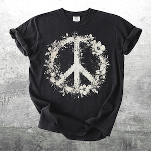May include: A black short-sleeve t-shirt featuring a large, distressed white graphic of a peace sign encircled by floral elements. The design has a weathered, textured appearance, and the shirt has a crew neckline and rolled sleeves.