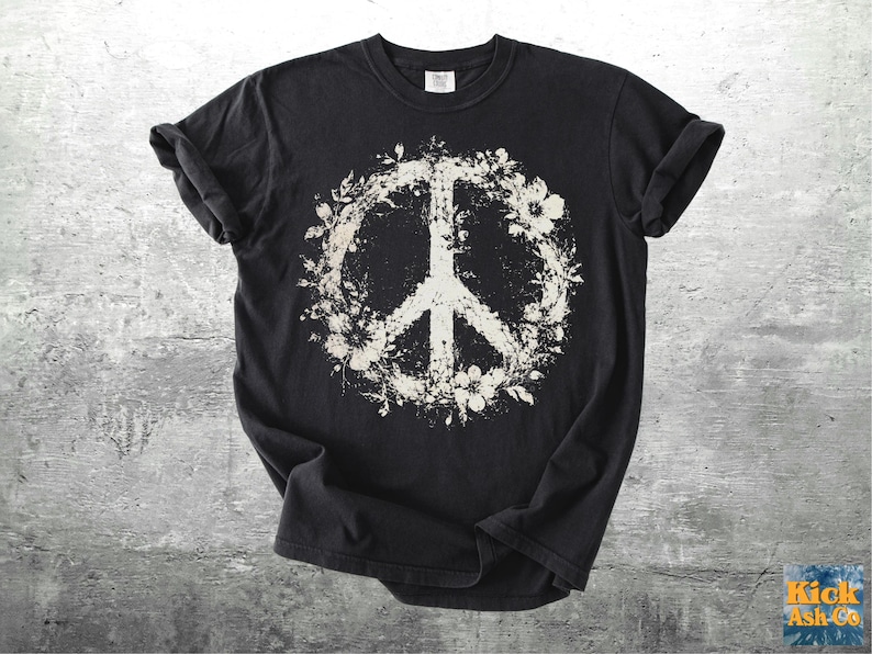 May include: A black short-sleeve t-shirt featuring a large, distressed white graphic of a peace sign encircled by floral elements. The design has a weathered, textured appearance, and the shirt has a crew neckline and rolled sleeves.