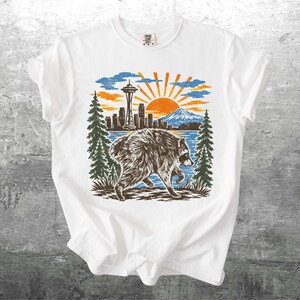 May include: A white short-sleeved t-shirt featuring a graphic print of a raccoon walking in front of a Seattle skyline, including the Space Needle, a mountain, pine trees, and a bright orange sun set against a blue sky background.