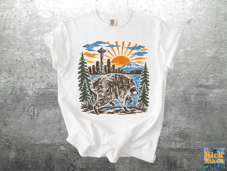 May include: A white short-sleeved t-shirt featuring a graphic print of a raccoon walking in front of a Seattle skyline, including the Space Needle, a mountain, pine trees, and a bright orange sun set against a blue sky background.