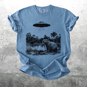 May include: A light blue t-shirt featuring a black and white graphic of a capybara standing near a body of water under a hovering UFO. The design includes palm trees and other foliage. The shirt has a crew neck and short sleeves.